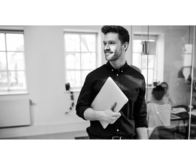 Smiling young businessman walking with his laptop in a modern office with colleagues at work in the background