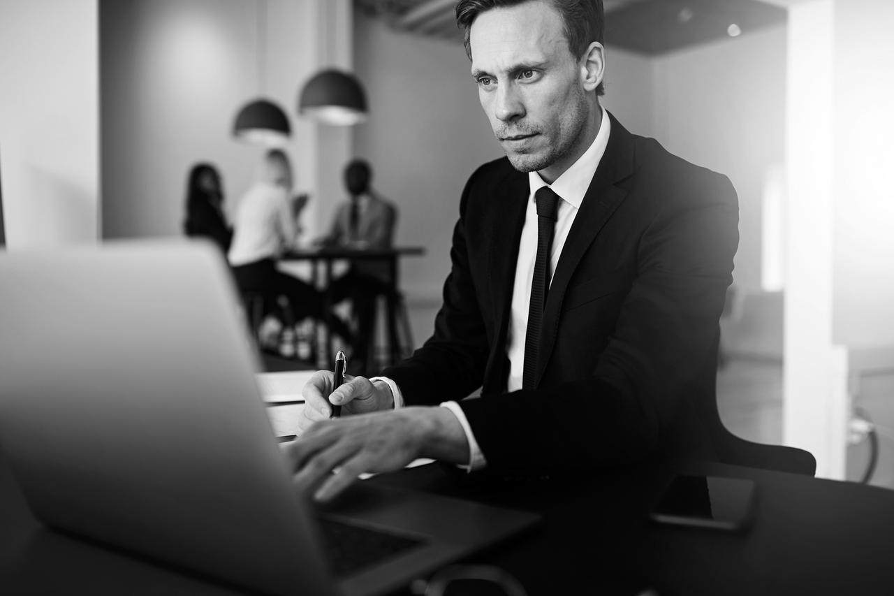 Businessman with laptop taking notes in office setting Businessman working online with a laptop while sitting at his desk in a bright modern office with colleagues in the background